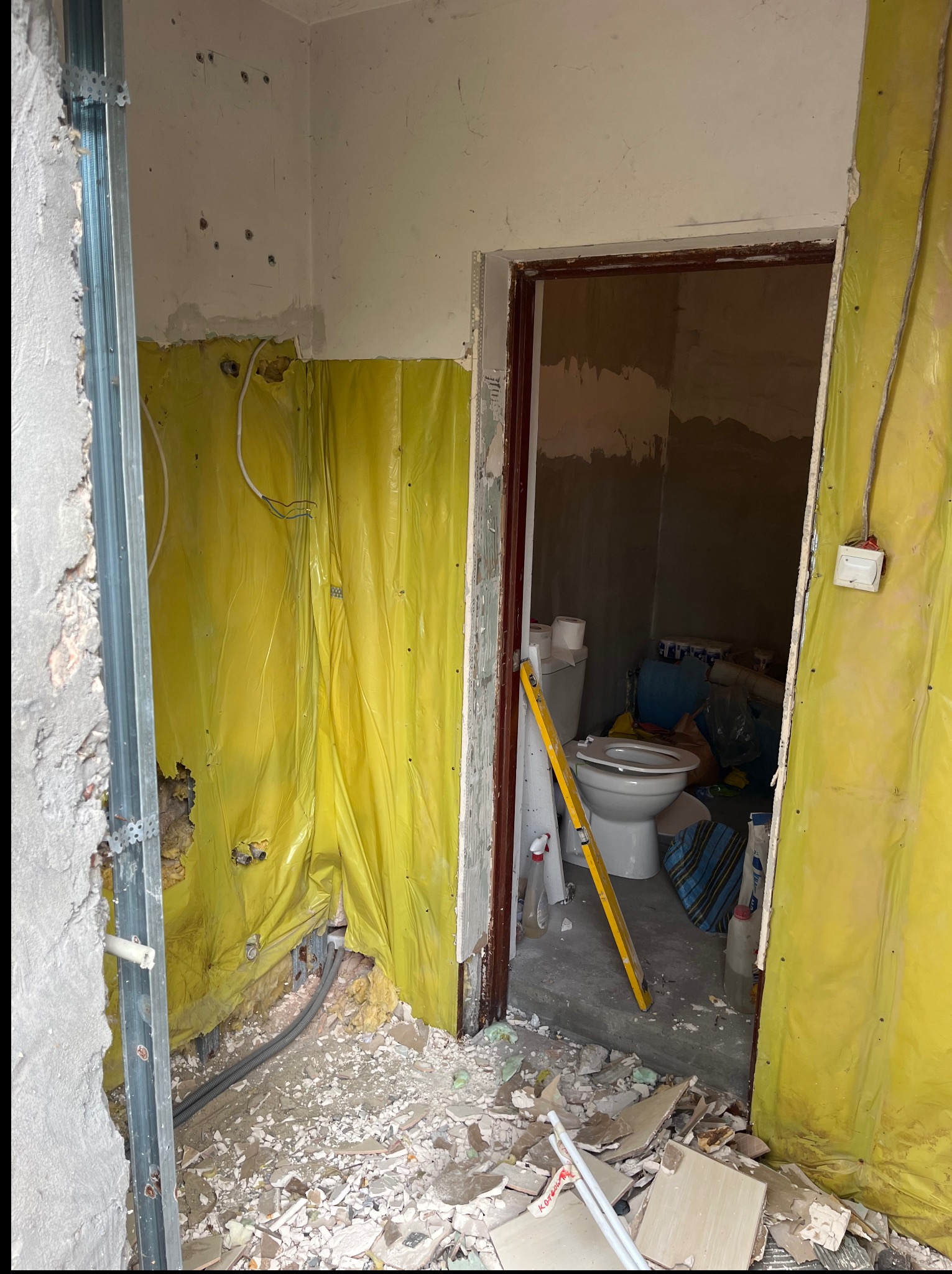 Wnętrze łazienki w trakcie remontu. Widoczne elementy konstrukcyjne, instalacje, zabezpieczenia folią i gruz. Poziomica oparta o toaletę. Ujęcie zza futryny.