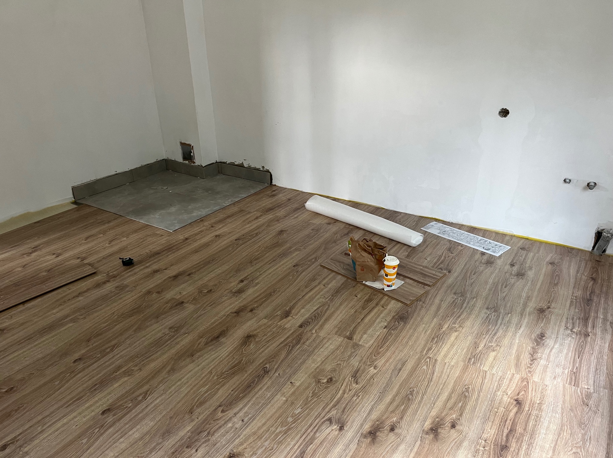 Układanie paneli podłogowych w jasnym wnętrzu. Widoczne narzędzia, rolka folii i niedokończona podłoga z imitacją drewna. Fragment ściany z widocznymi instalacjami.