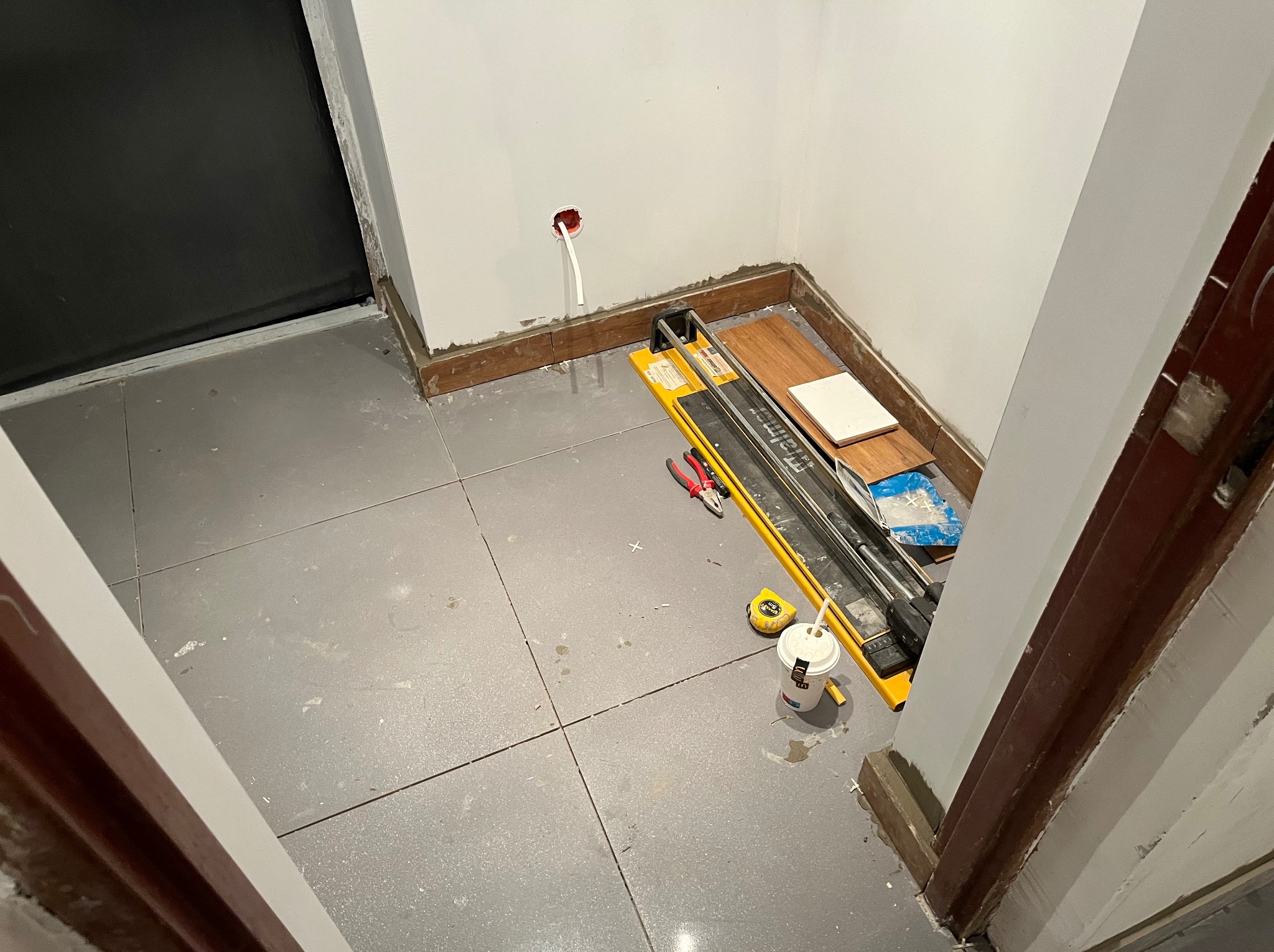 Szare płytki podłogowe w trakcie układania, widoczny żółty przecinak do płytek, miarka i puszka. Fragment ściany z gniazdkiem i listwą przypodłogową.