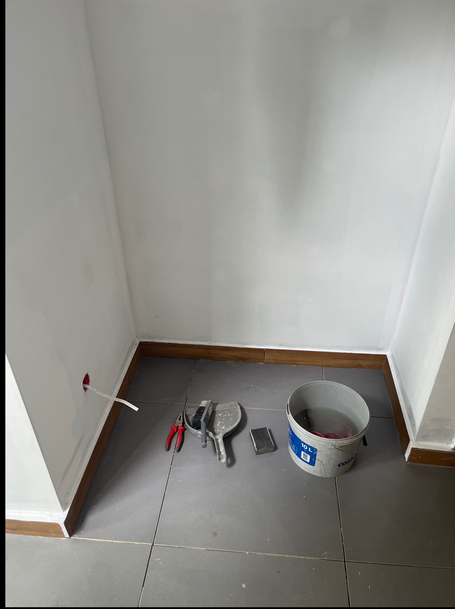 Wnętrze w trakcie odświeżania: białe ściany z widocznym gniazdkiem, szare płytki podłogowe, wiadro z wodą i narzędzia malarskie. Listwa przypodłogowa w kolorze drewna.