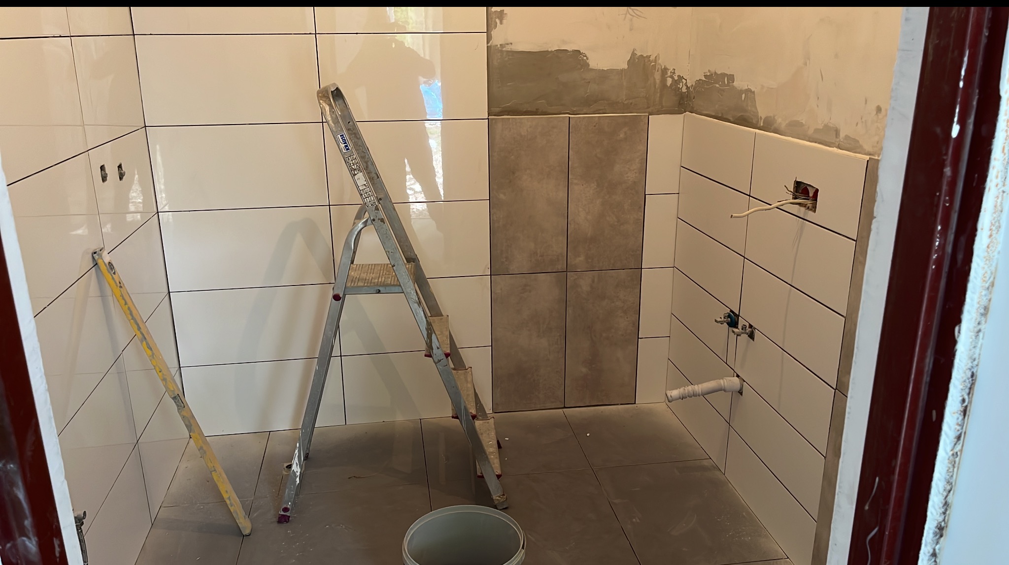 Układanie białych, prostokątnych płytek w łazience, widoczna drabina i poziomica, szare płytki w centralnej części ściany, instalacja wodna i elektryczna.