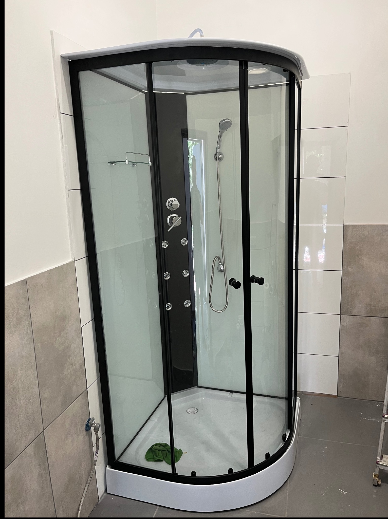 Kabina prysznicowa z czarnymi profilami i hydromasażem, zamontowana w wykończonej łazience z szarymi i białymi płytkami. Widoczny fragment drabiny.