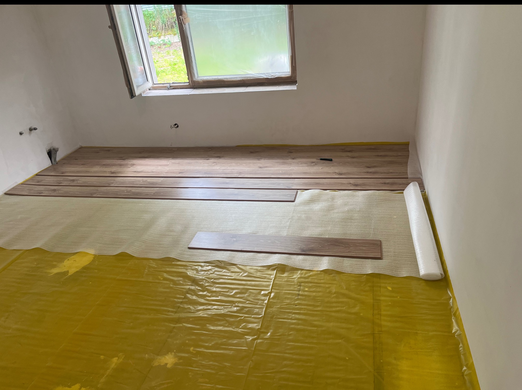 Układanie paneli podłogowych w pokoju na żółtej folii. Częściowo ułożone panele, widoczne okno dachowe i fragment ściany. Etap prac wykończeniowych.
