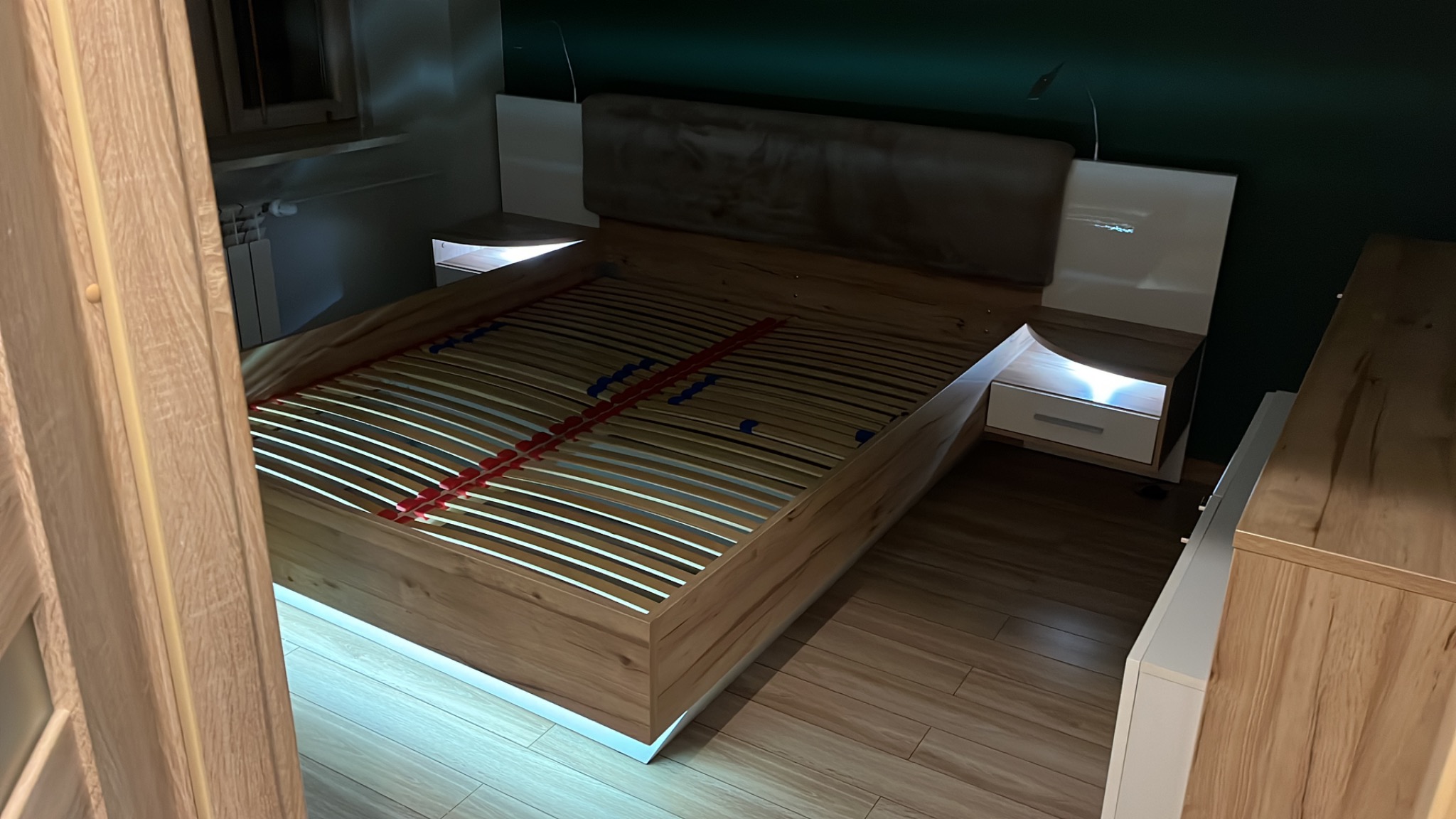 Drewniane łóżko z podświetleniem LED i stelażem listwowym, widoczne z perspektywy pokoju, z elementami szafek nocnych i wezgłowia. Ustawione na panelach podłogowych.