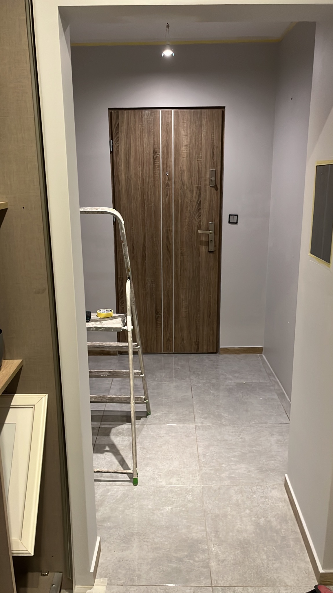 Korytarz w trakcie odświeżania: drabina malarska, zabezpieczone taśmą krawędzie sufitu i drzwi. Widoczne płytki podłogowe i świeżo pomalowane ściany w odcieniach szarości.
