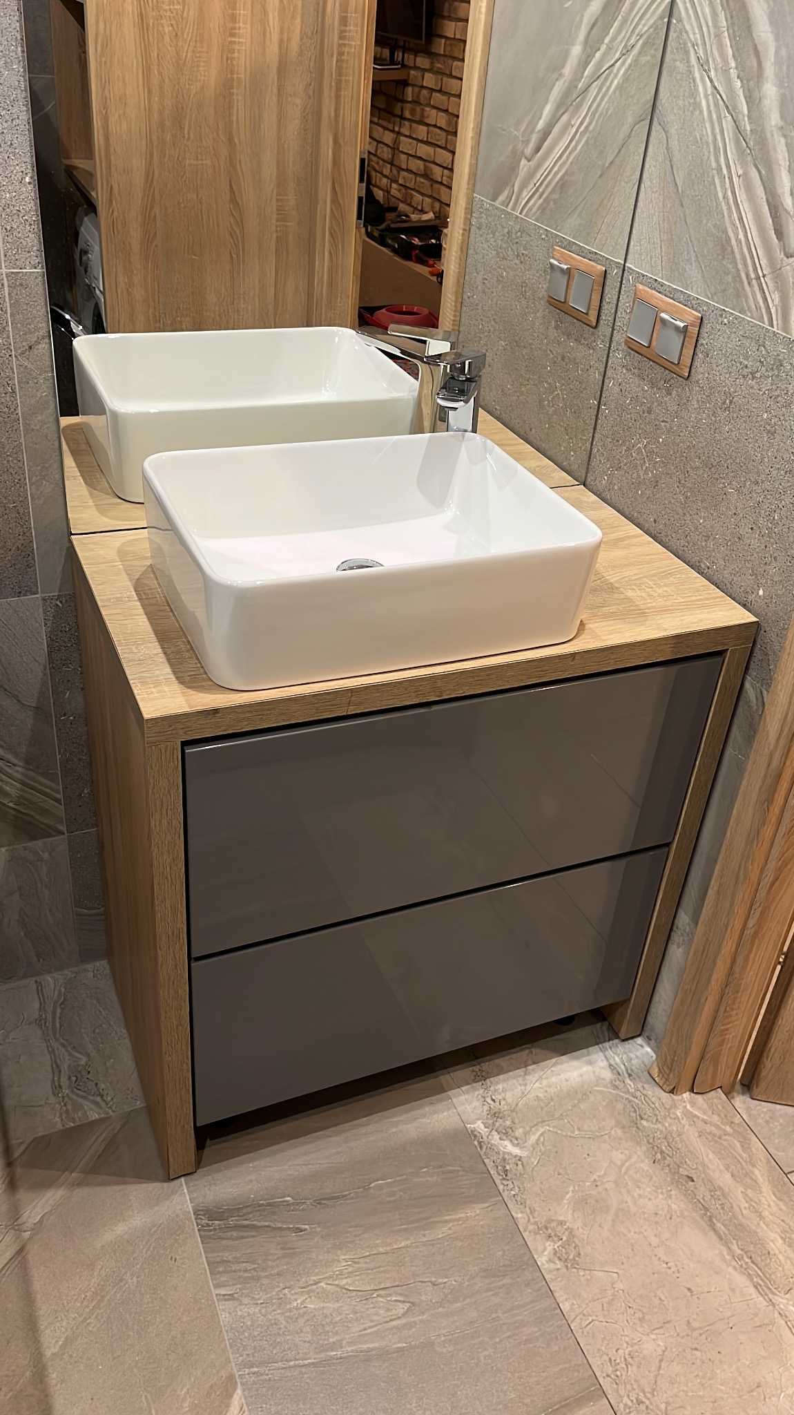 Nowoczesna łazienka: biała, prostokątna umywalka nablatowa na szafce z szarymi frontami, drewnopodobny blat i płytki imitujące kamień na ścianie i podłodze.