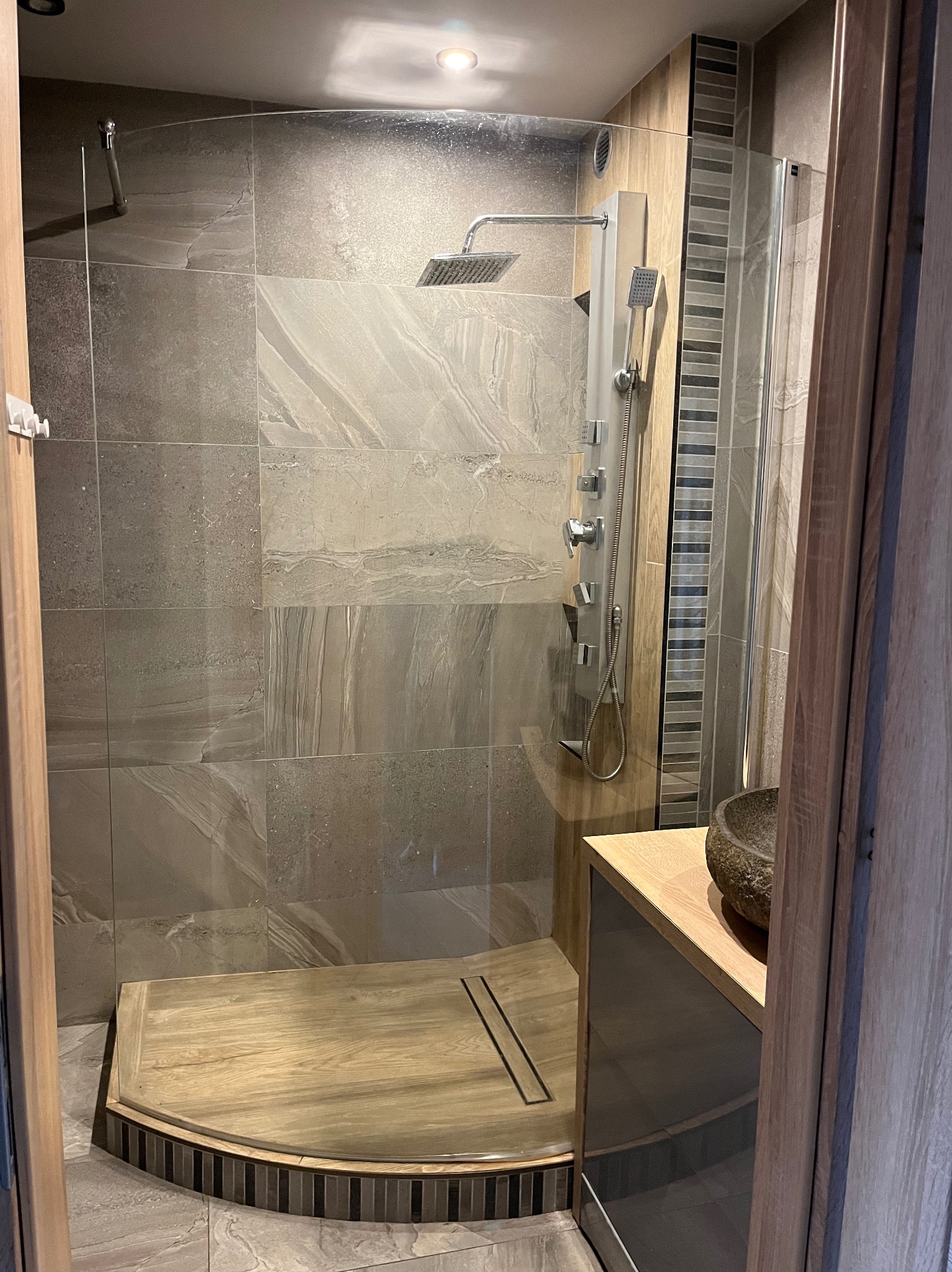 Nowoczesna kabina prysznicowa z drewnianym brodzikiem i panelami imitującymi kamień. Szklana ściana oddziela strefę umywalki z kamienną misą. Minimalistyczny design.