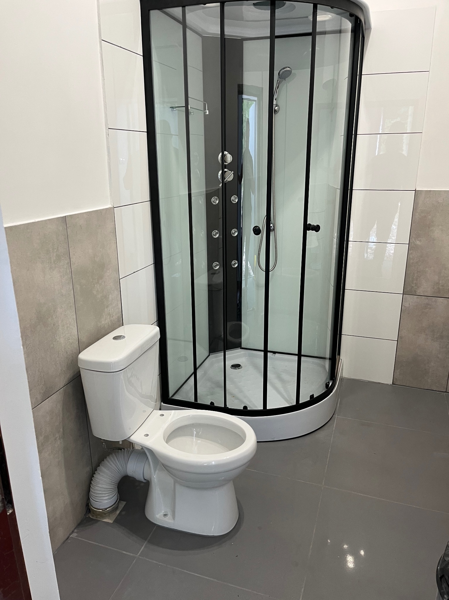 Nowoczesna kabina prysznicowa z czarną ramą i biała toaleta w wykończonej łazience z szarymi płytkami podłogowymi i częściowo wyłożonymi ścianami.