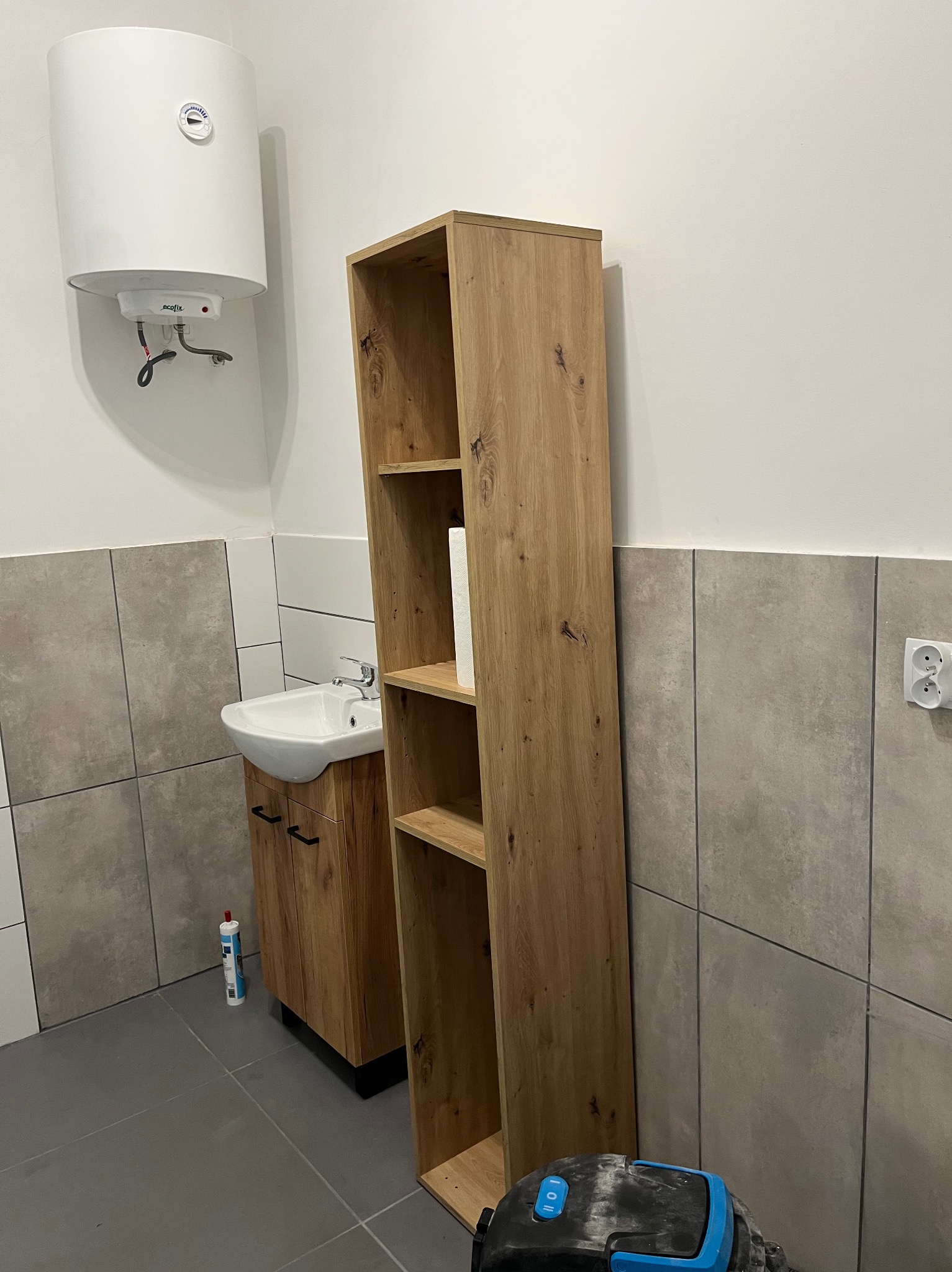 Wykończona łazienka z drewnianą szafką pod umywalką, wysokim regałem i bojlerem na ścianie. Szare płytki na ścianach i podłodze. Widoczny odkurzacz.