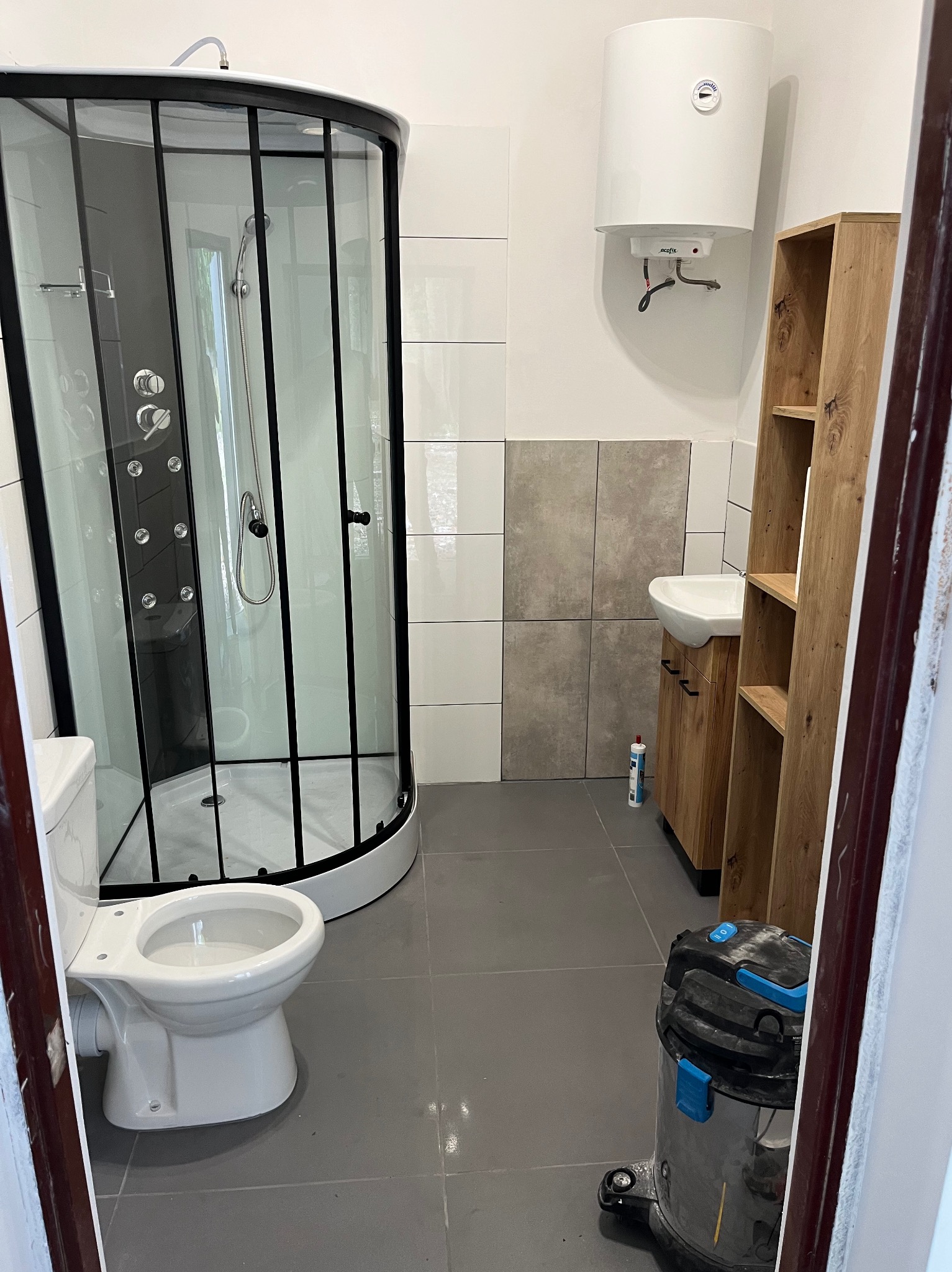 Nowoczesna łazienka z kabiną prysznicową typu walk-in z czarnymi ramami, umywalką z szafką z drewna i szarymi płytkami podłogowymi. Widoczny bojler i odkurzacz.