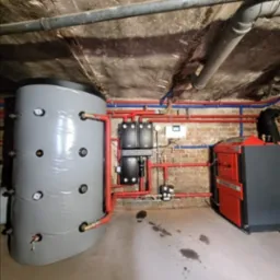 Kotłownia z piecem Atmos, zbiornikiem akumulacyjnym i instalacją grzewczą z rur miedzianych. Widoczne elementy sterujące i zabezpieczające. Ściany z cegły, sufit betonowy.