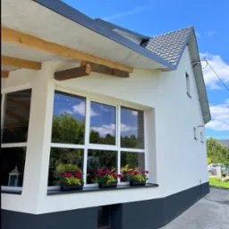 Biała elewacja domu z szarym cokołem i dachem, okna z kwiatami w doniczkach, błękitne niebo w tle. Widoczny fragment elewacji bocznej z oknami.