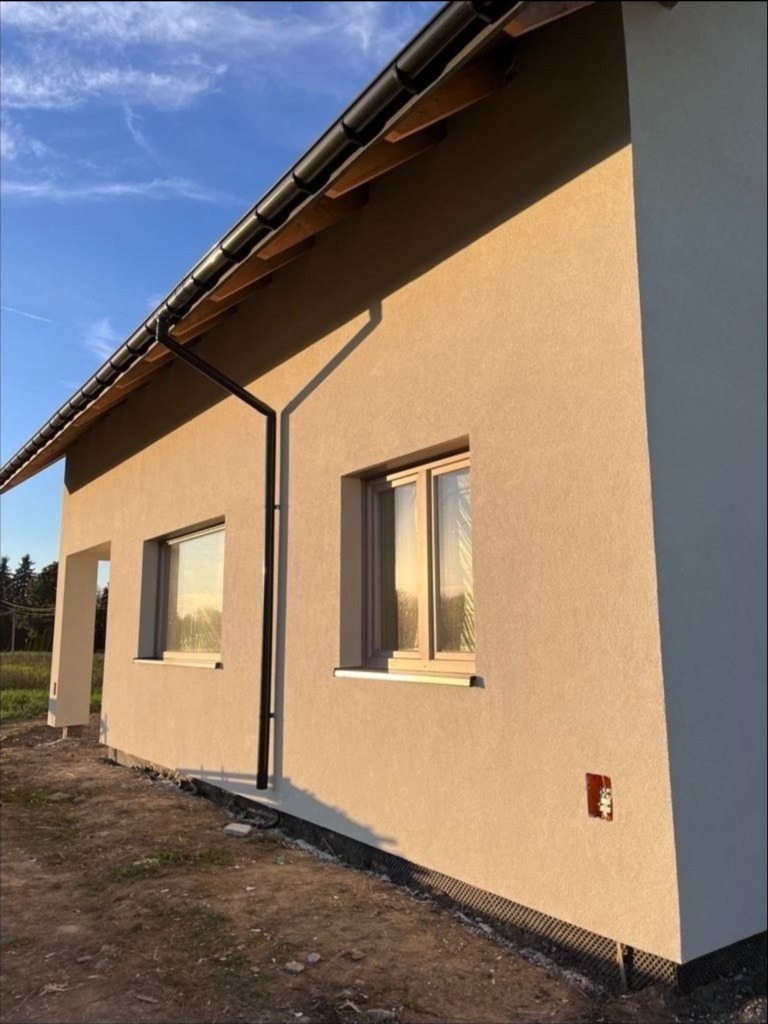 Jasny, otynkowany dom jednorodzinny z ciemną rynną, widziany pod kątem w promieniach zachodzącego słońca. Widoczne okna i fragment terenu.