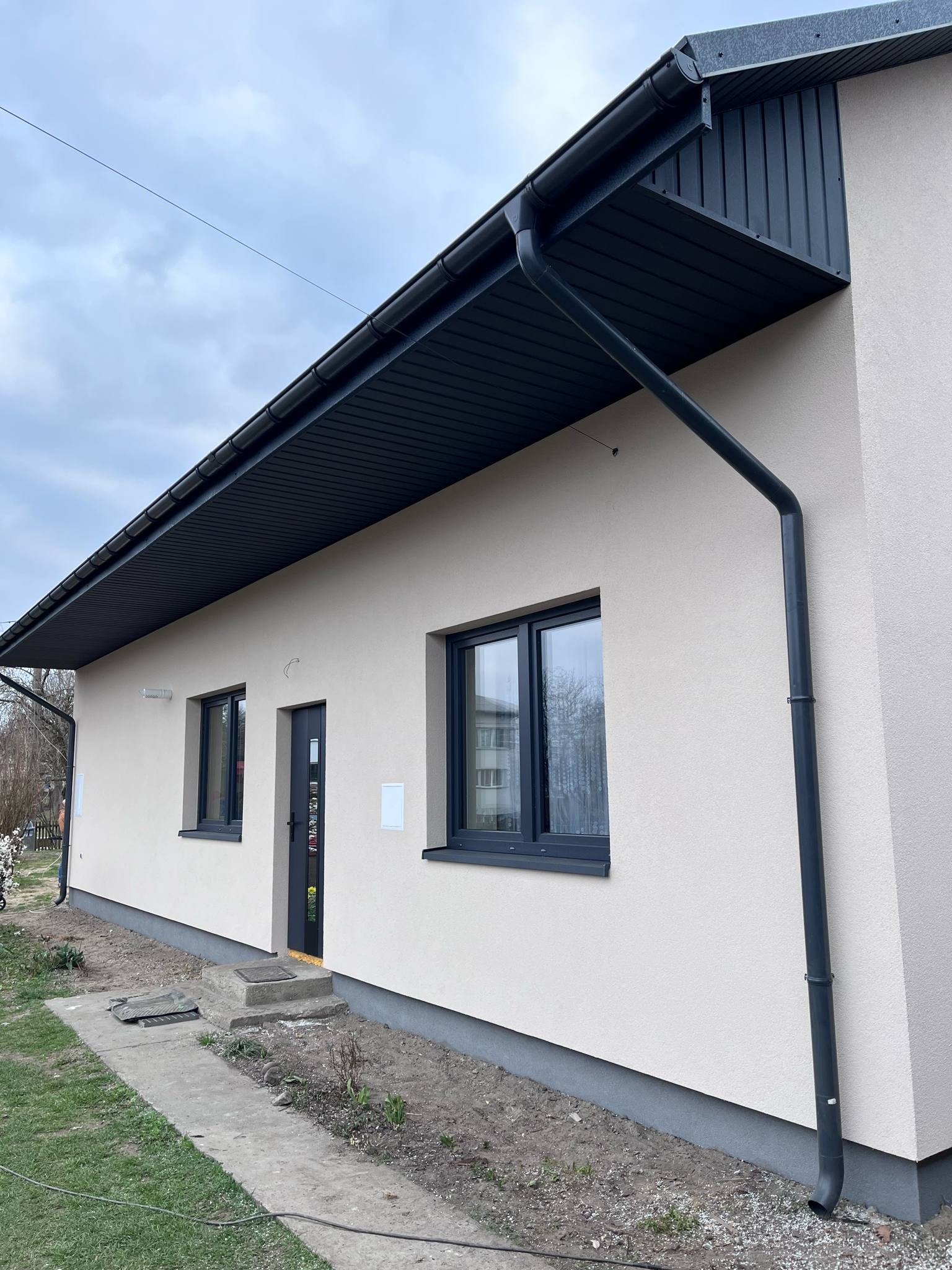 Jasna elewacja domu jednorodzinnego z ciemnymi oknami i orynnowaniem. Widoczne fragmenty zieleni i chodnika. Dom po renowacji z nową elewacją.