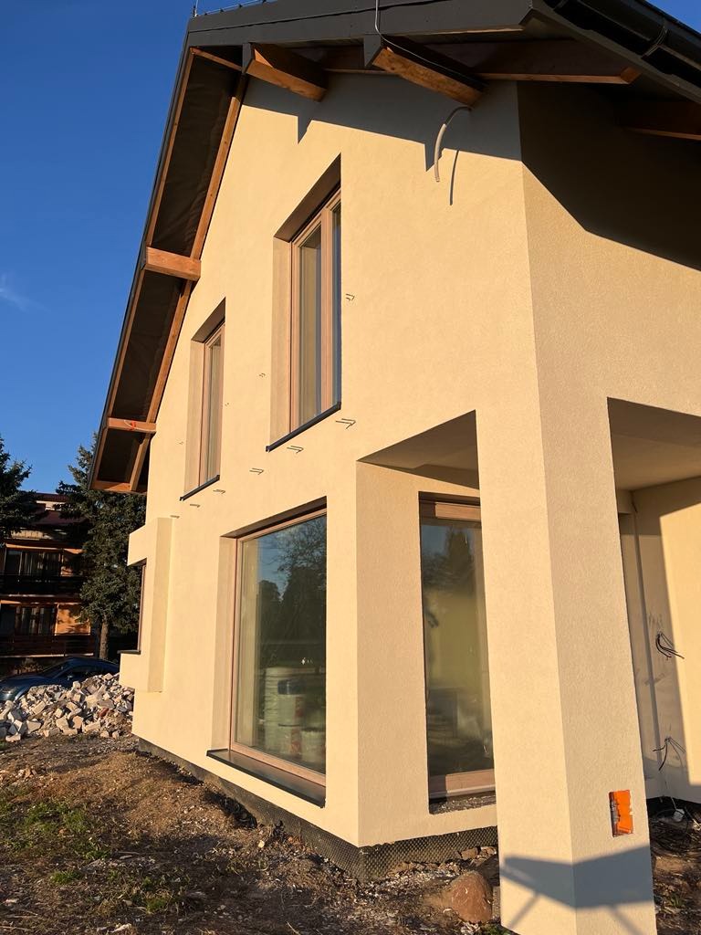 Jasna, otynkowana elewacja domu jednorodzinnego z dużymi oknami, widoczna w promieniach zachodzącego słońca. Uwagę zwraca minimalistyczny styl i nowoczesne wykończenie.