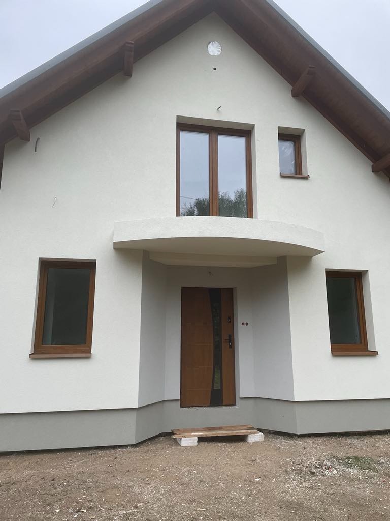 Biała elewacja domu jednorodzinnego z drewnianymi oknami i drzwiami, widok na wejście główne z małym daszkiem, szara podmurówka, teren wokół domu niezagospodarowany.