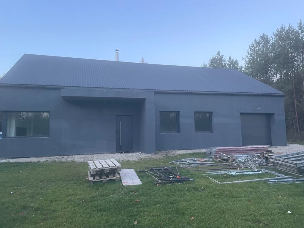 Nowoczesny dom z szarą elewacją i blachodachówką na tle lasu. Widoczne rusztowania i palety, co sugeruje prace wykończeniowe.