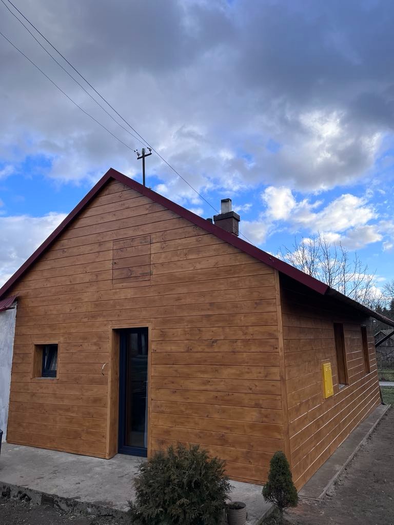 Dom z elewacją z drewnianych paneli w ciepłym odcieniu brązu, widoczny fragment dachu i komin na tle pochmurnego, błękitnego nieba.