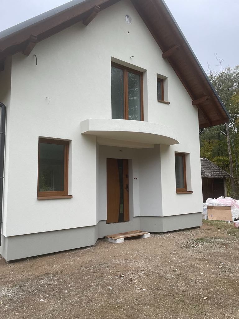 Dom jednorodzinny z jasną elewacją i drewnianą stolarką okienną. Widoczny fragment dachu i otoczenie budynku. Świeżo położony tynk.