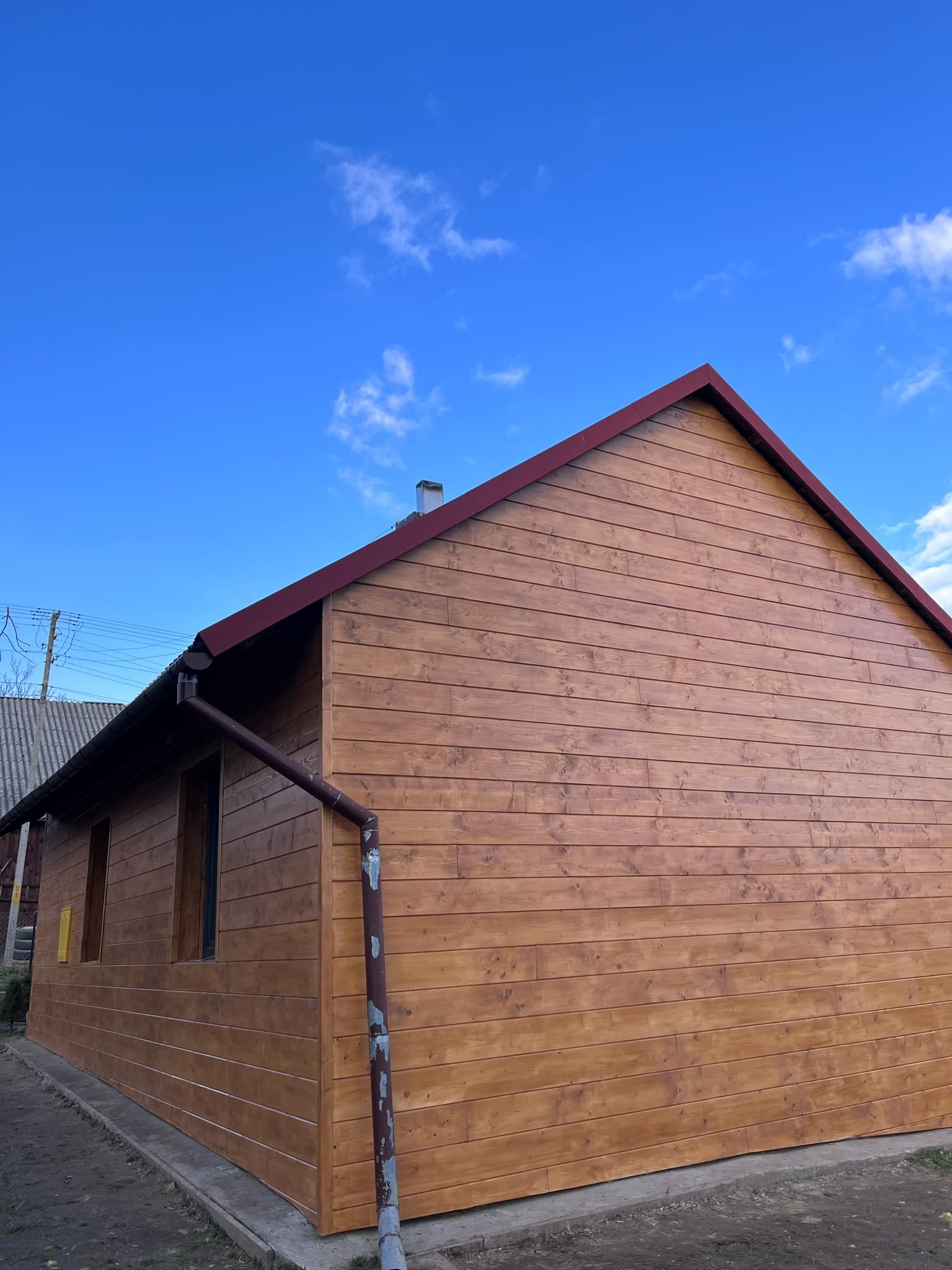 Dom z elewacją z drewnianych paneli w ciepłym, brązowym odcieniu, widoczny fragment rynny i czerwone obramowanie dachu na tle błękitnego nieba.