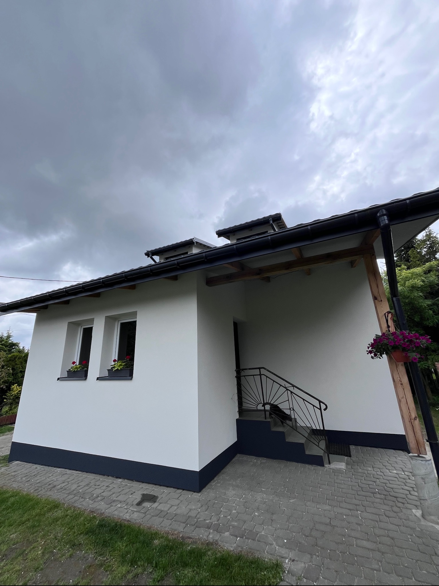 Biała elewacja domu jednorodzinnego z ciemną podmurówką, widok z dołu, pochmurne niebo w tle. Zadbany ogród i kostka brukowa.