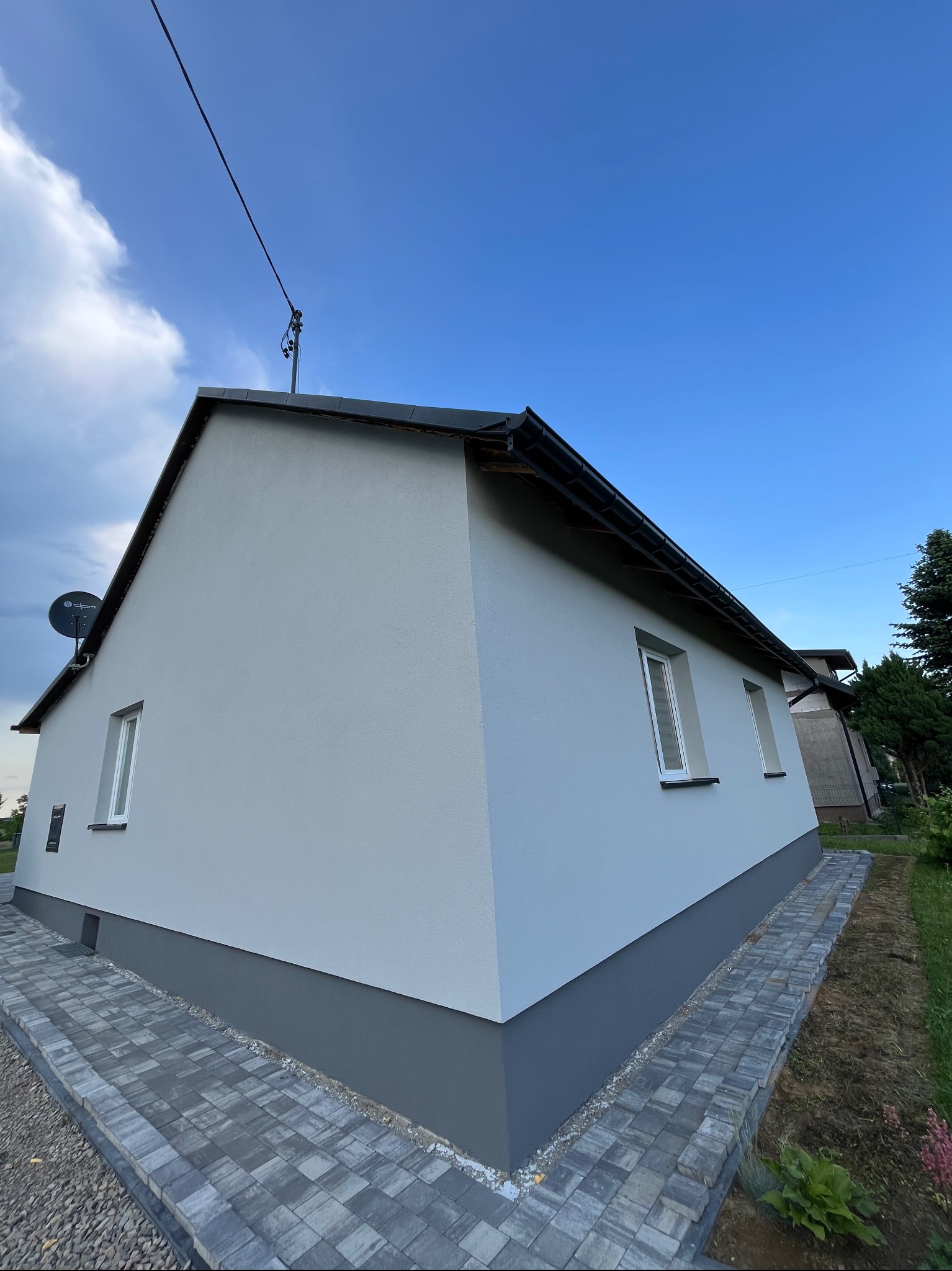 Jasnoszary dom jednorodzinny po renowacji elewacji, z nową kostką brukową wokół. Widoczna antena satelitarna i fragment ogrodu. Ujęcie z dołu.