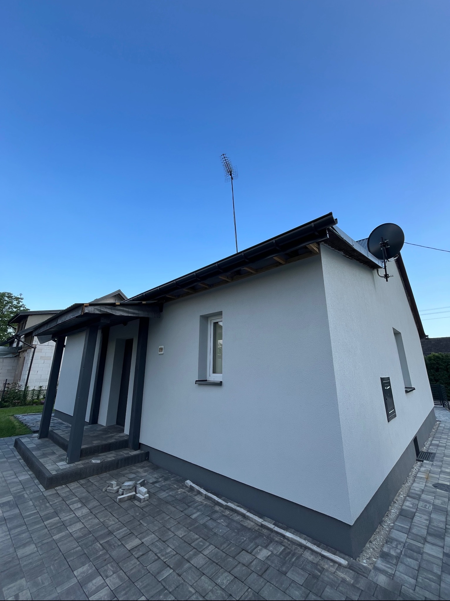 Szary, jednopiętrowy dom z ciemnymi akcentami, widziany pod kątem. Widoczne elementy wykończenia elewacji i chodnika z kostki brukowej. Antena na dachu.