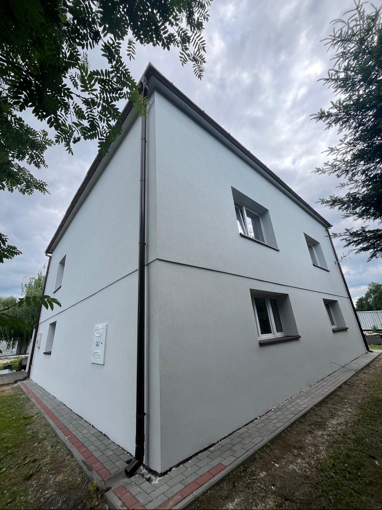 Dwukondygnacyjny dom z jasną, świeżo wykończoną elewacją, widoczny pod kątem. Na ścianie skrzynka gazowa. Ciemna rynna. W tle drzewa i pochmurne niebo.
