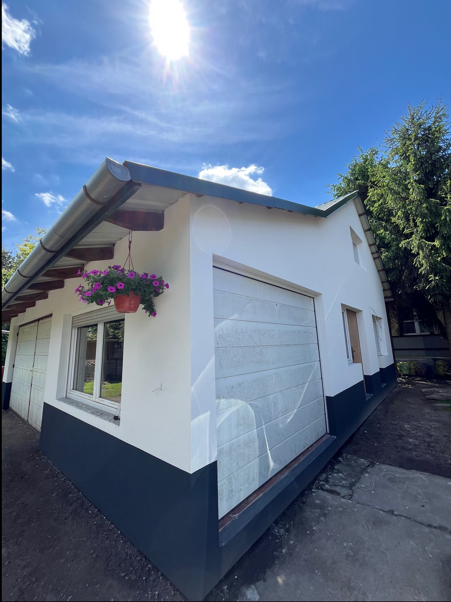 Biała elewacja budynku z garażem, z ciemnym cokołem. Wisząca donica z kwiatami przy oknie. Słoneczny dzień, błękitne niebo. Widok z dołu.