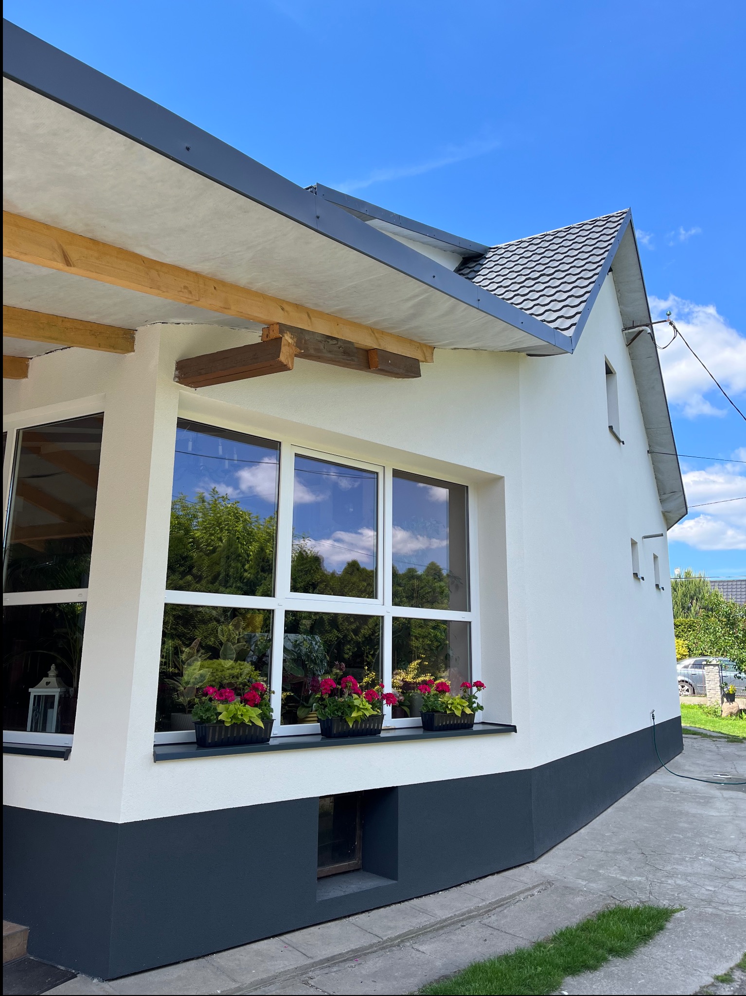 Biała elewacja domu z szarym cokołem i dachem, okna z kwiatami w doniczkach, błękitne niebo w tle. Widoczny fragment elewacji bocznej z oknami.