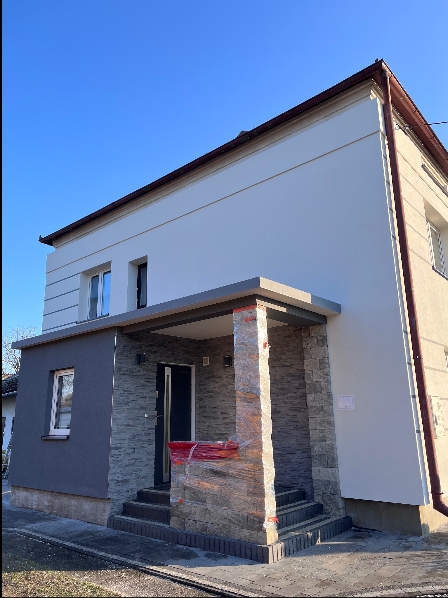 Nowoczesny dom po renowacji elewacji z elementami kamiennymi, kolumny wejściowe zabezpieczone folią. Jasne ściany, grafitowe detale i błękitne niebo w tle.