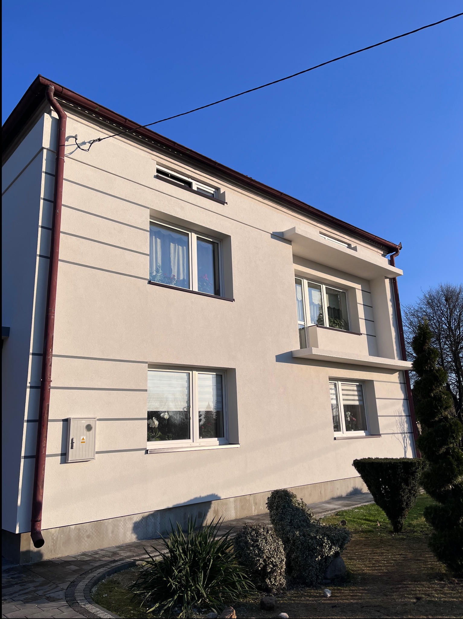 Dwupiętrowy dom z kremową elewacją, brązowymi rynnami i dekoracyjnymi pasami. Widok z dołu ukazuje okna z kwiatami i zadbany ogród.