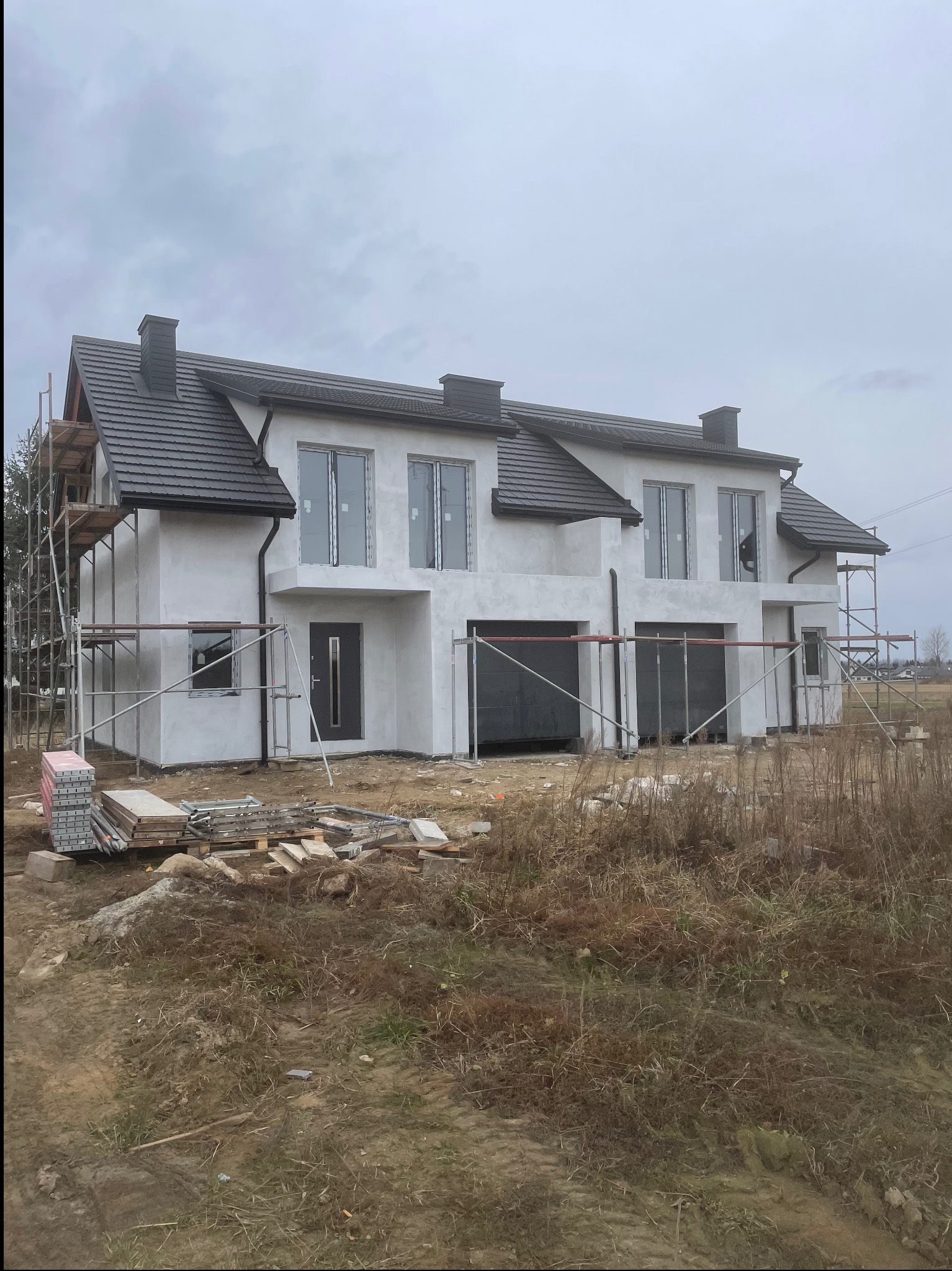 Surowy stan domu bliźniaczego w Pruchnik podczas prac elewacyjnych. Widoczne rusztowania, okna i drzwi. Elewacja w trakcie tynkowania, przygotowana do malowania.