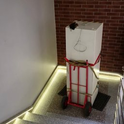 Artur Pieróg - Biały prostopadłościan zabezpieczony pasami na czerwonym wózku transportowym, stoi na schodach z ledowym oświetleniem i ceglanym tłem.