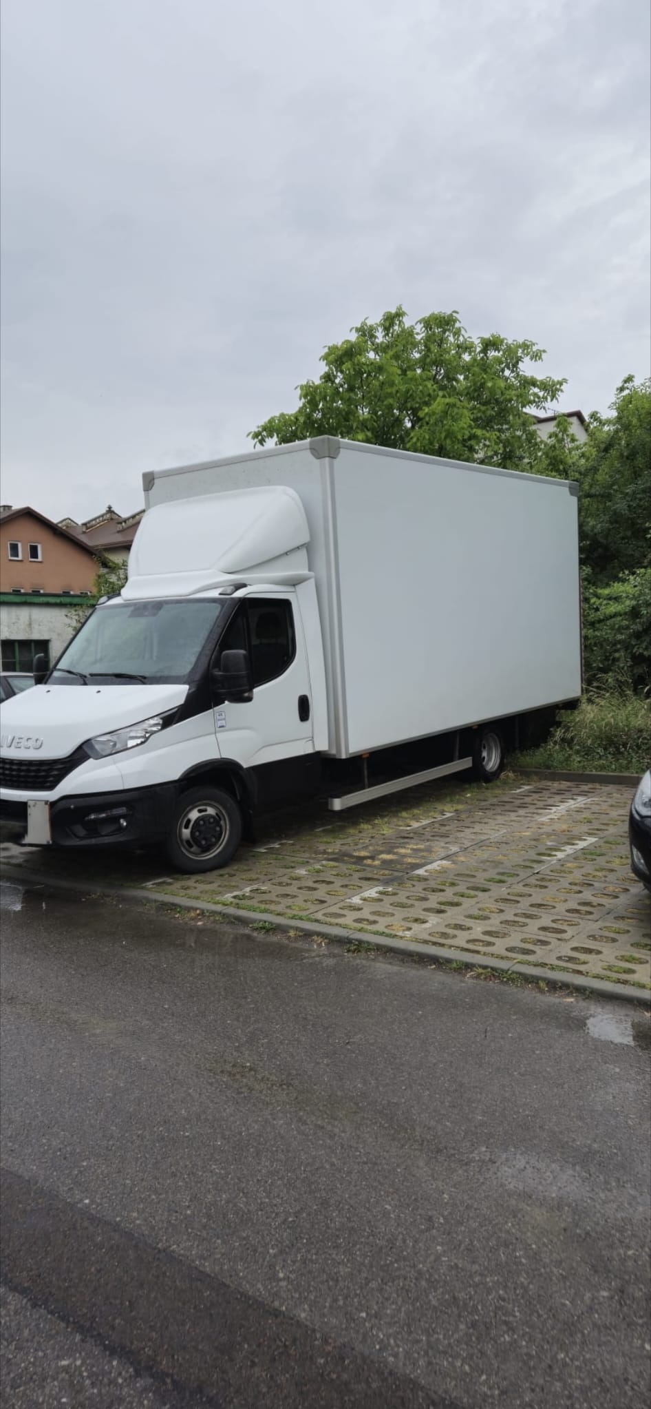 Biały samochód dostawczy Iveco z zabudową kontenerową zaparkowany na betonowej nawierzchni. W tle zieleń i budynki mieszkalne. Pochmurne niebo.