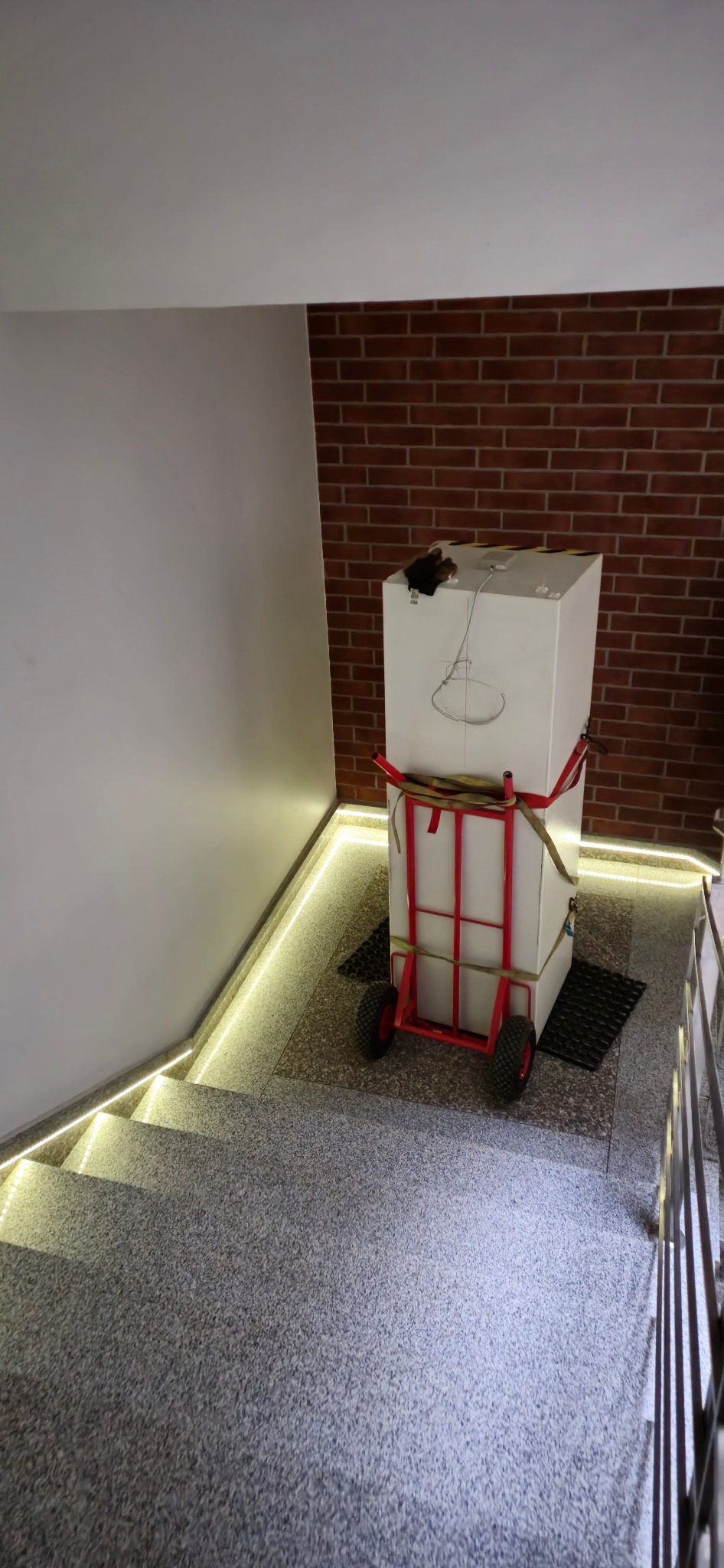 Biały prostopadłościan zabezpieczony pasami na czerwonym wózku transportowym, stoi na schodach z ledowym oświetleniem i ceglanym tłem.