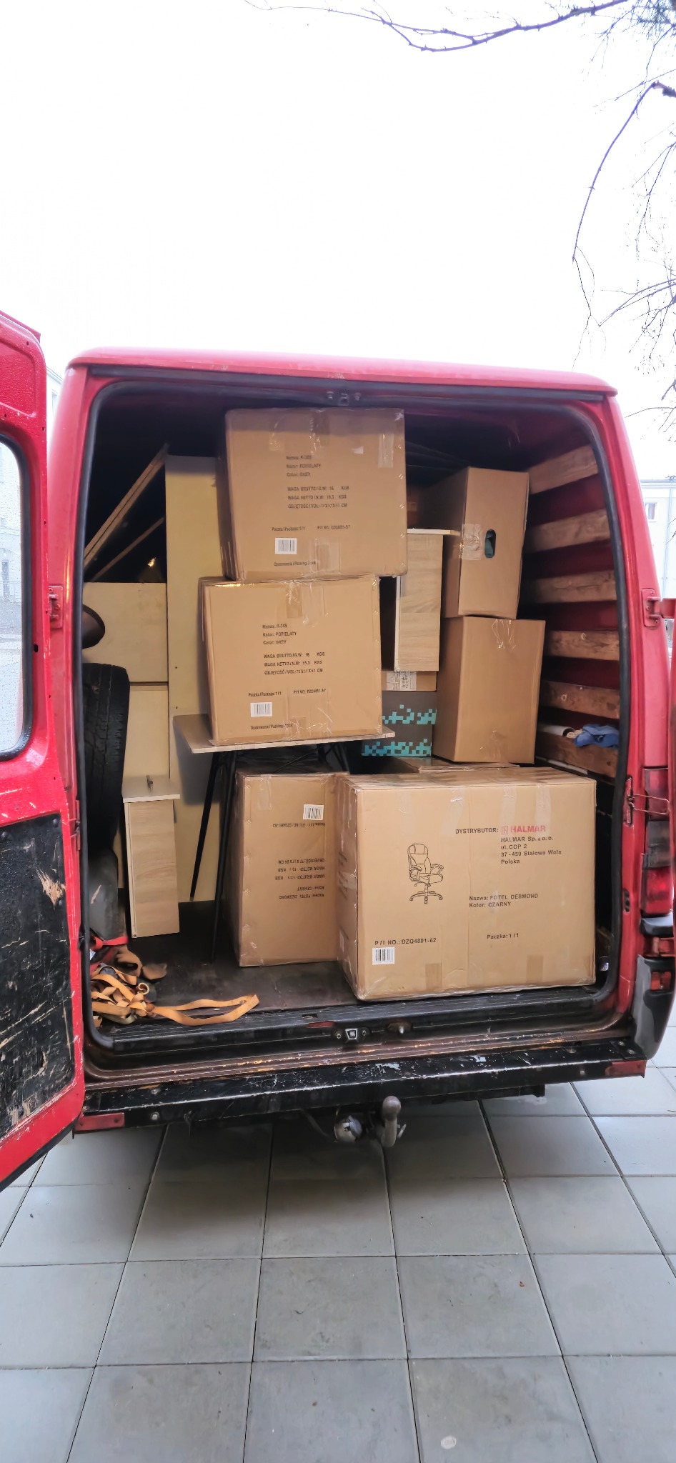 Czerwony van z otwartymi drzwiami bagażnika, wypełniony kartonami i meblami gotowymi do transportu. Widoczne zabezpieczenia ładunku i częściowo koło zapasowe.
