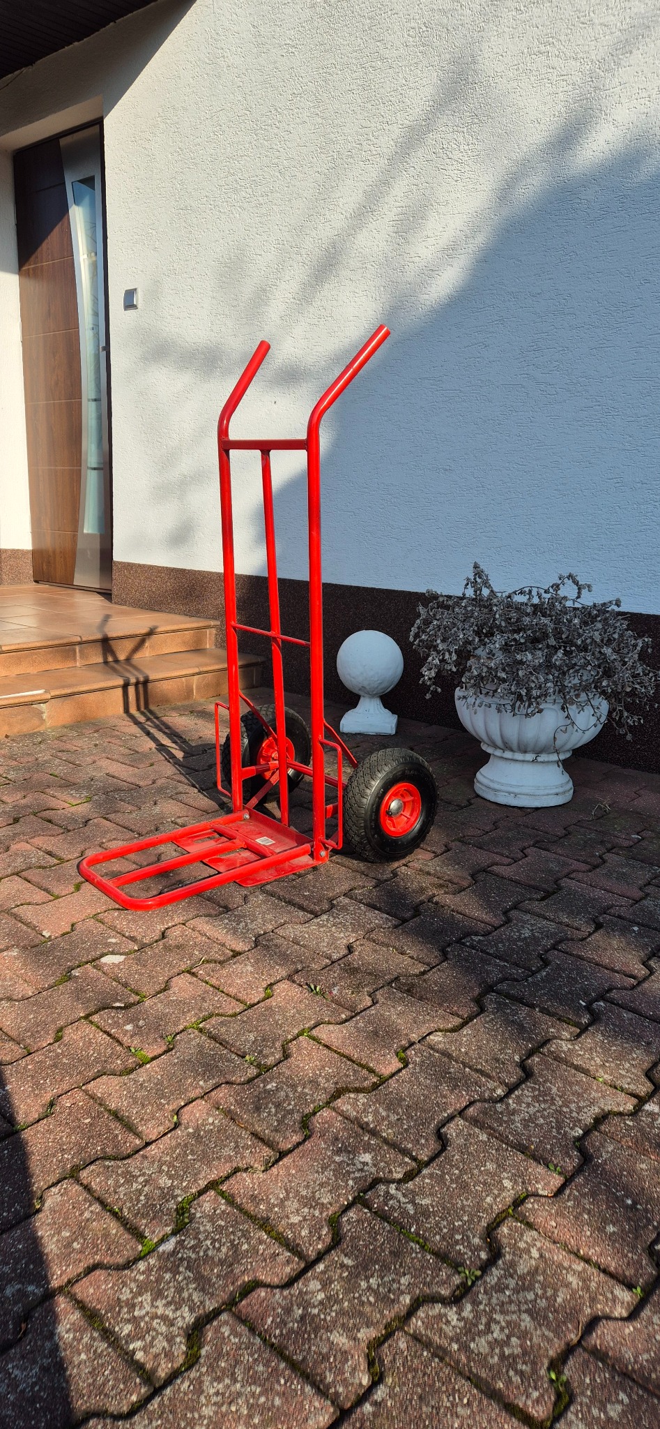 Czerwony wózek transportowy na kostce brukowej przed budynkiem z dekoracyjnymi elementami. Wózek gotowy do użycia, oświetlony naturalnym światłem.