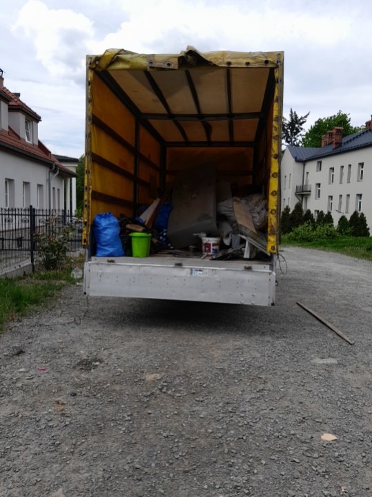 Kontener transportowy wypełniony przedmiotami, widok z tyłu, sugerujący opróżnianie mieszkania lub transport po remoncie. Budynki w tle.