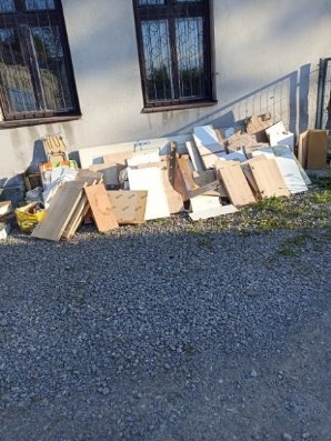 Sterta odpadów budowlanych (płyty, kartony) leżąca na żwirze przed budynkiem z oknami w kratkę. Odpady po remoncie.
