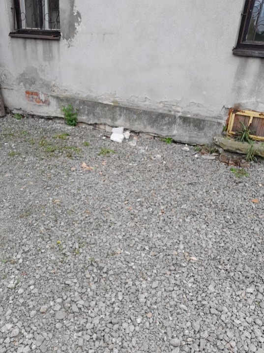 Fragment elewacji budynku z widocznymi uszkodzeniami i gruzem na żwirowym podłożu. Widoczne okno i fragment muru.