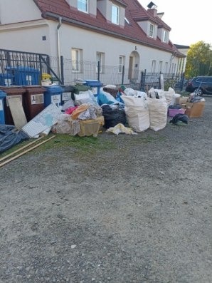 Opróżnianie posesji: sterta odpadów i worków na żwirze przed budynkiem mieszkalnym, obok widoczne pojemniki na śmieci i fragment ogrodzenia.