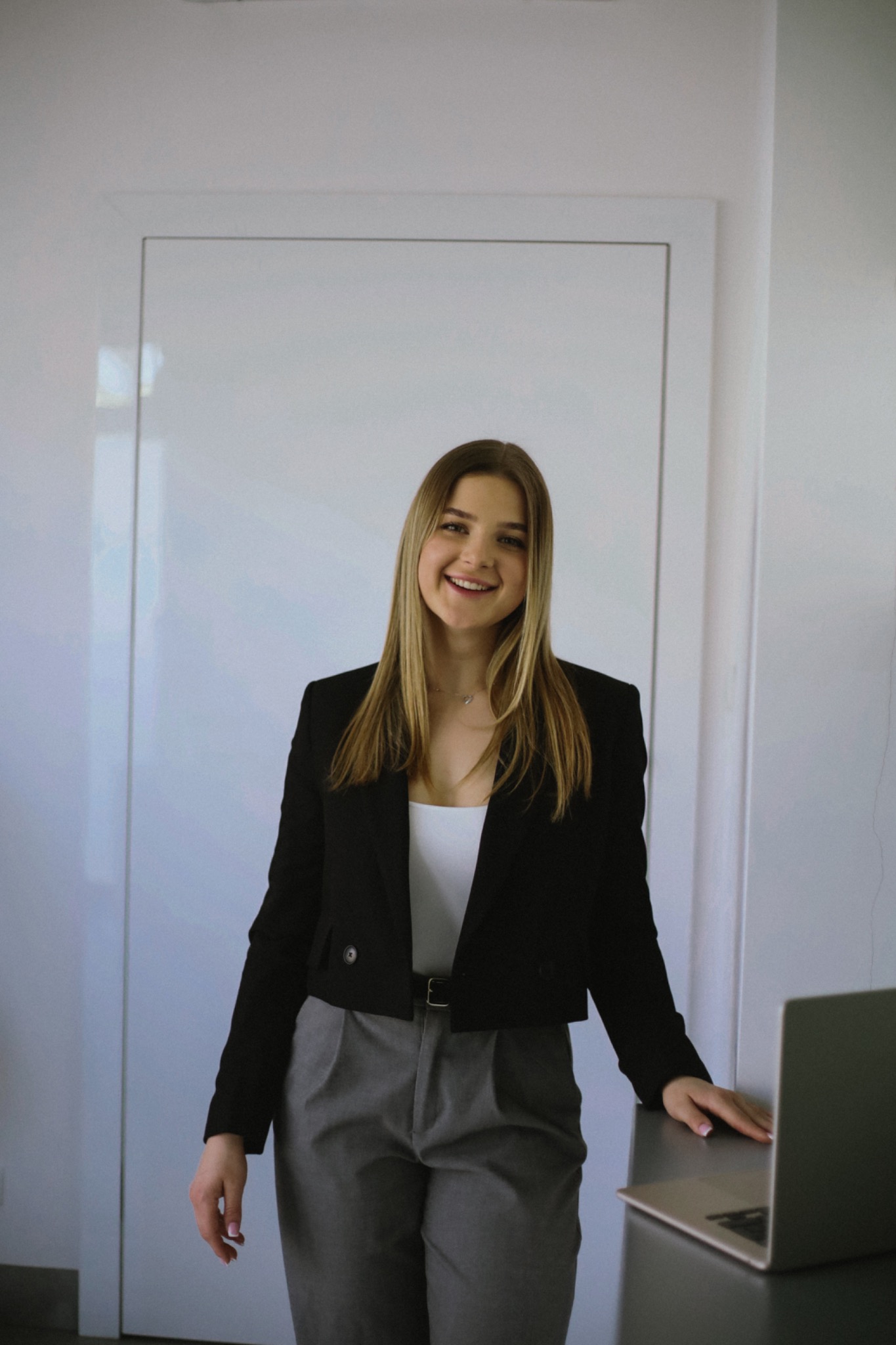 Uśmiechnięta młoda kobieta w czarnej marynarce i szarych spodniach, stojąca obok laptopa, na tle białej ściany. Portret dietetyka w gabinecie.