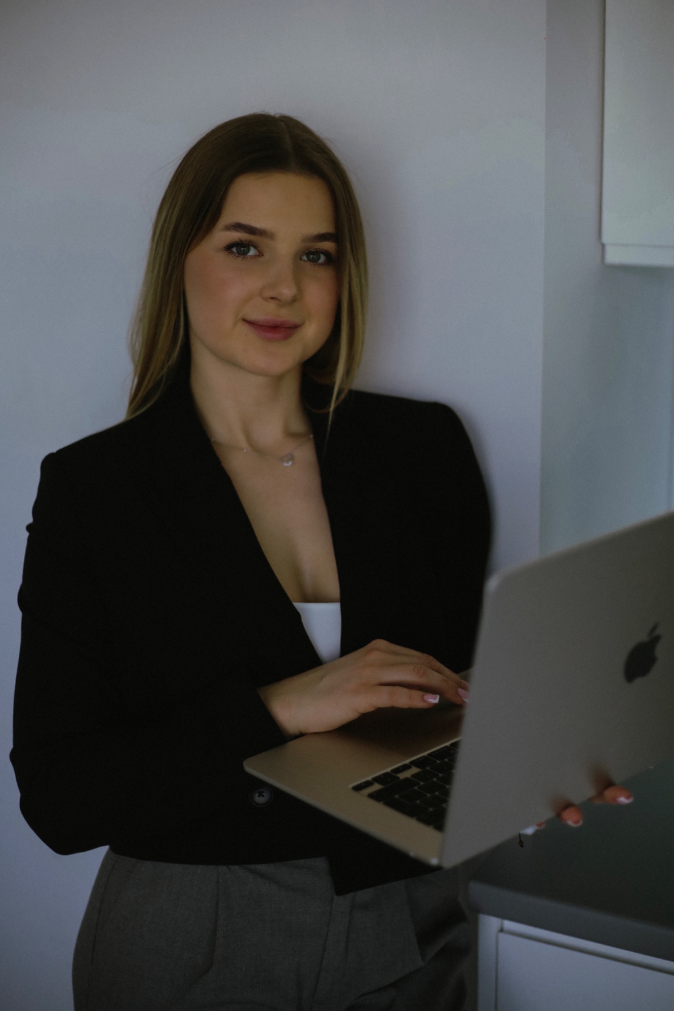 Młoda kobieta w czarnej marynarce, z delikatnym uśmiechem, trzymająca laptopa w jasnym wnętrzu. Widoczny fragment blatu i szafki.