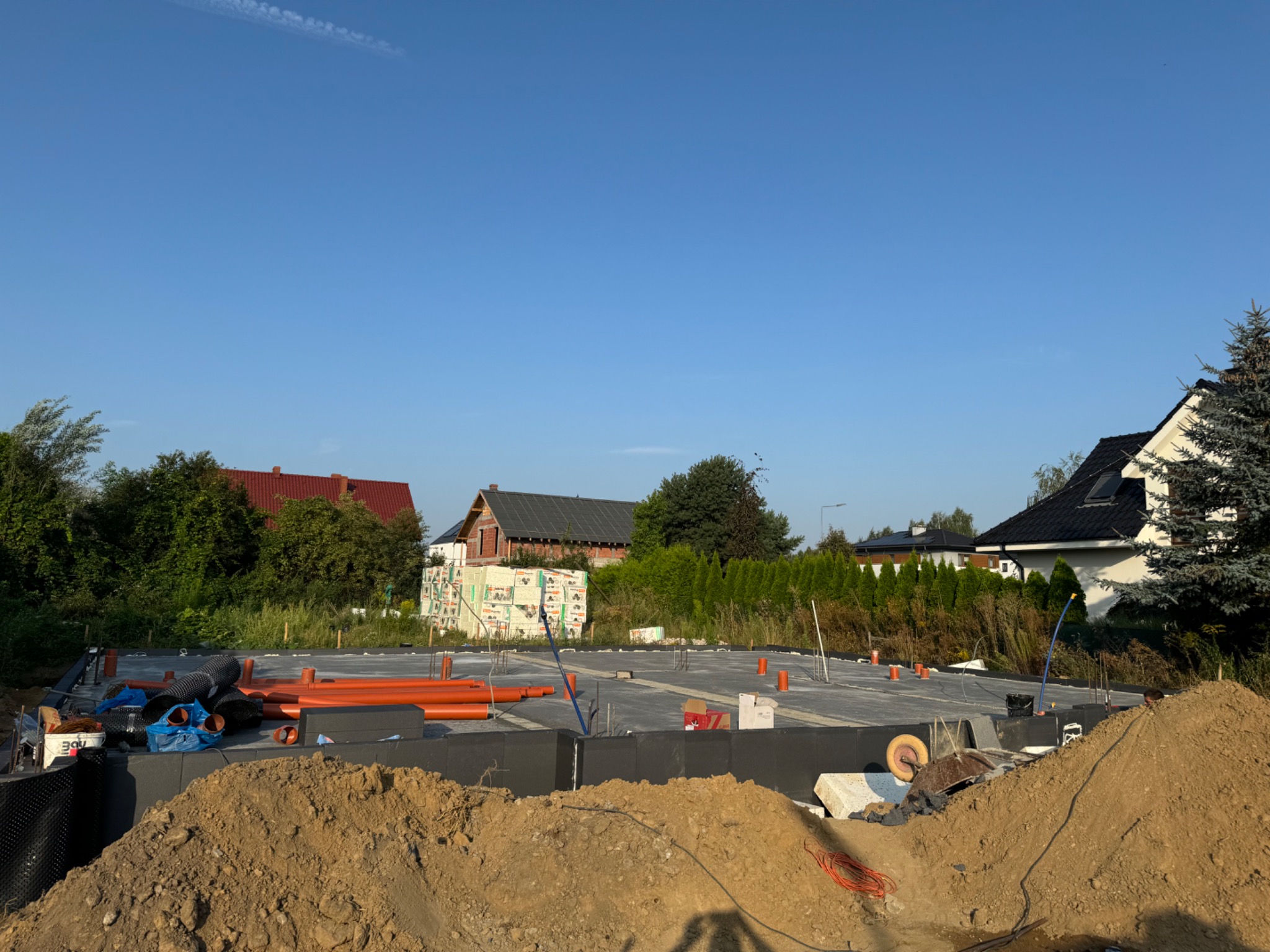 Fundamenty domu w trakcie budowy: izolacja, rury i zbrojenie na tle błękitnego nieba i zabudowy jednorodzinnej w słoneczny dzień. Widoczny nasyp ziemi.