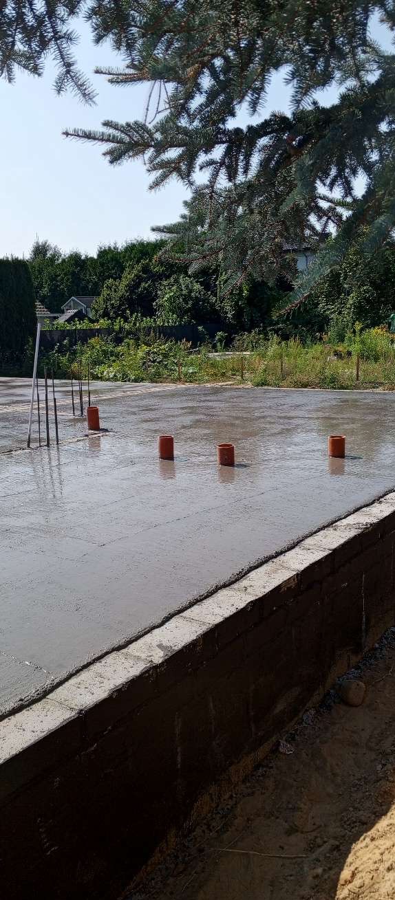 Zalany fundament z wystającymi rurami i prętami, widok z bliska, w tle zieleń i błękitne niebo. Ujęcie w dziennym świetle, widoczna struktura betonu i ziemi.