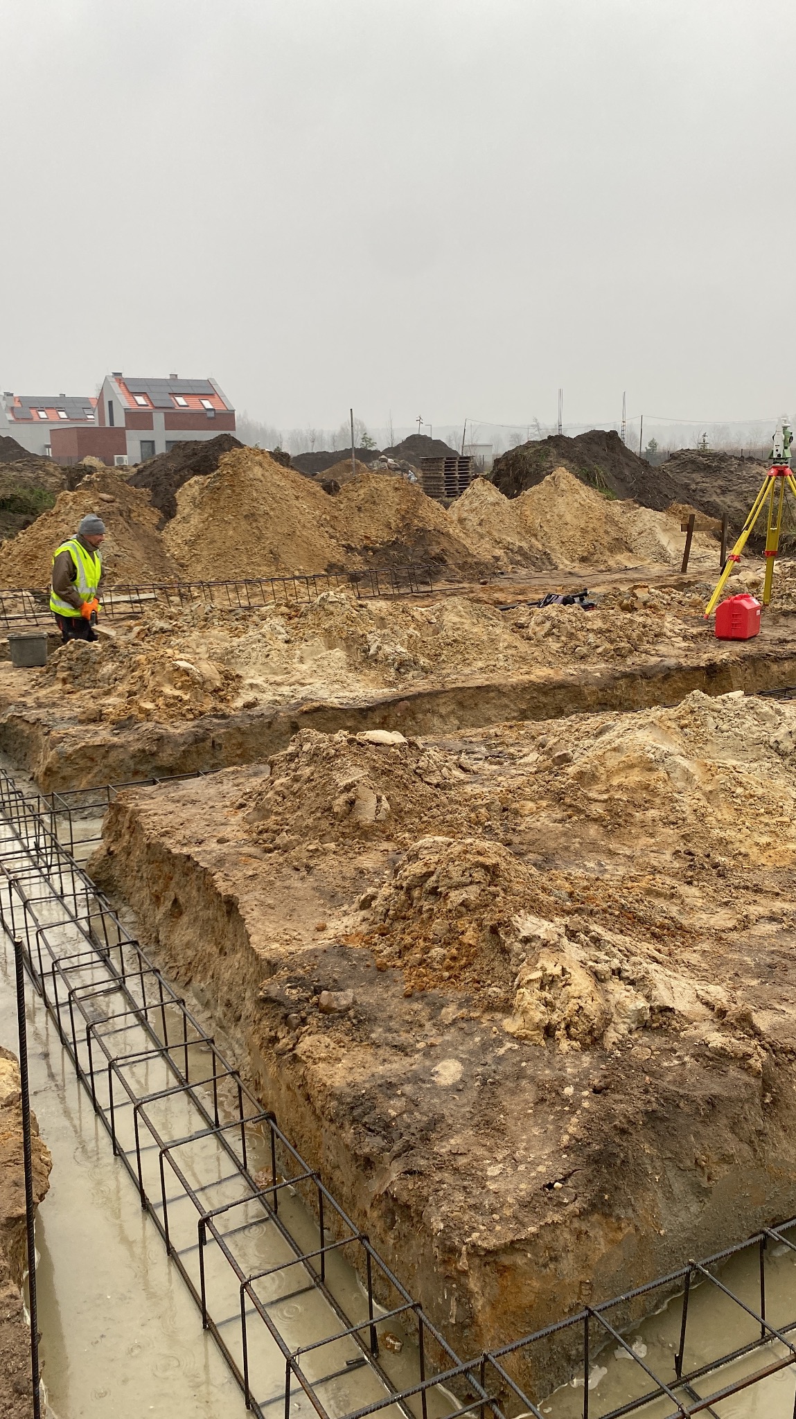 Wykopane rowy pod fundamenty z widocznym zbrojeniem i stertami ziemi w tle, geodeta w odblaskowej kamizelce i niwelator na trójnogu. Prace ziemne w pochmurny dzień.