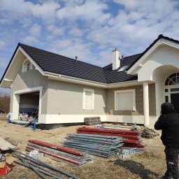 ONIX - Dom jednorodzinny w trakcie ocieplania, widoczne płyty izolacyjne z kołkami. Na pierwszym planie rusztowania i materiały budowlane. Pracownik w ubraniu roboczym.