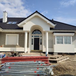 ONIX - Dom w trakcie prac elewacyjnych: widoczna warstwa ocieplenia, przygotowane rusztowania, czarne dachówki, biała stolarka okienna i kolumny wejściowe.