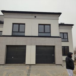 ONIX - Dwukondygnacyjny dom z dwoma garażami i jasną elewacją, widok z zewnątrz. Osoba w kurtce po prawej stronie. Kostka brukowa przed budynkiem, palety w tle.